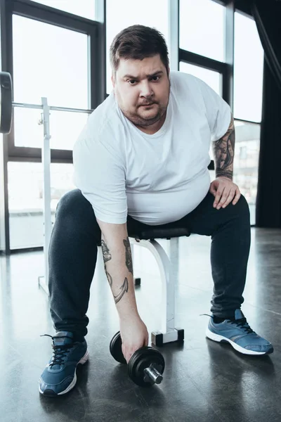 Hombre tatuado con sobrepeso mirando a la cámara y haciendo ejercicio con mancuerna en el centro deportivo - foto de stock