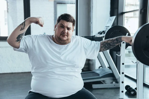 Overweight tattooed man sitting on bench, looking at camera and showing muscles at gym — Stock Photo