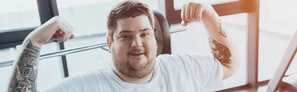 Panoramic shot of overweight tattooed man smiling and showing muscles at gym with sunlight — Stock Photo