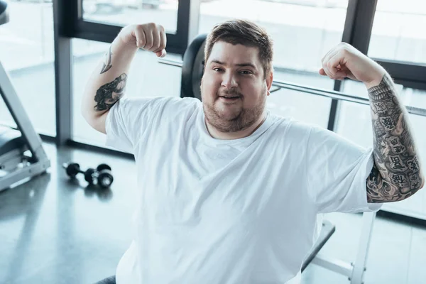 Overweight tattooed man smiling, looking at camera and showing muscles at gym — Stock Photo