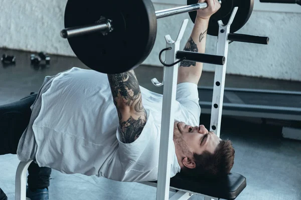 Hombre tatuado con sobrepeso en camiseta blanca haciendo ejercicio con barra en el gimnasio - foto de stock