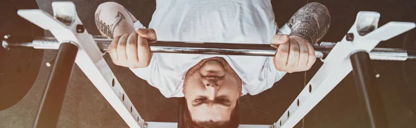 Panoramaaufnahme eines übergewichtigen tätowierten Mannes beim Training mit der Langhantel im Fitnessstudio mit Sonnenlicht — Stockfoto