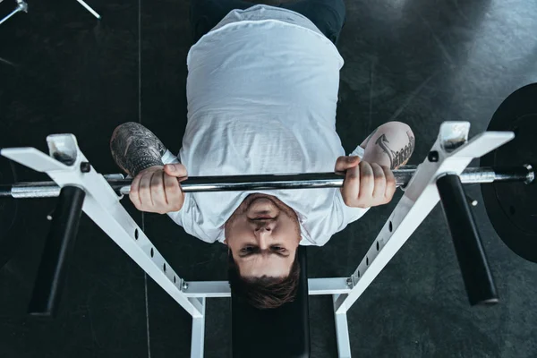Вид зверху на татуйованого чоловіка з надмірною вагою в білій футболці тренування з барбелом у спортзалі — стокове фото