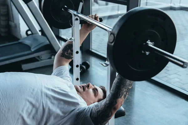 Sobrepeso hombre tatuado entrenamiento con barra de pesas en el gimnasio - foto de stock