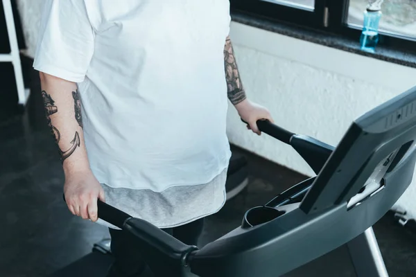 Частичный вид человека с татуировкой с избыточным весом, бегущего на беговой дорожке в спорткомплексе — стоковое фото