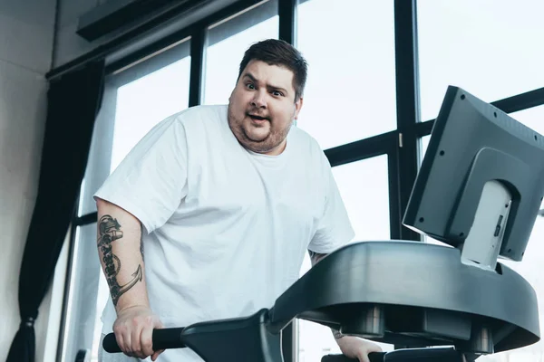 Übergewichtiger tätowierter Mann blickt in Kamera und läuft im Sportzentrum auf Laufband — Stockfoto
