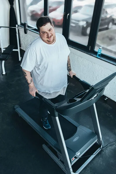 Smiling overweight tattooed man looking at camera while running on treadmill at sports center — Stock Photo
