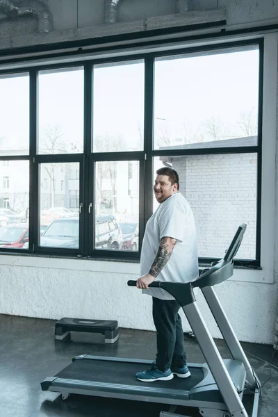 Übergewichtiger tätowierter Mann blickt beim Training auf Laufband im Sportzentrum mit Kopierraum in die Kamera — Stockfoto