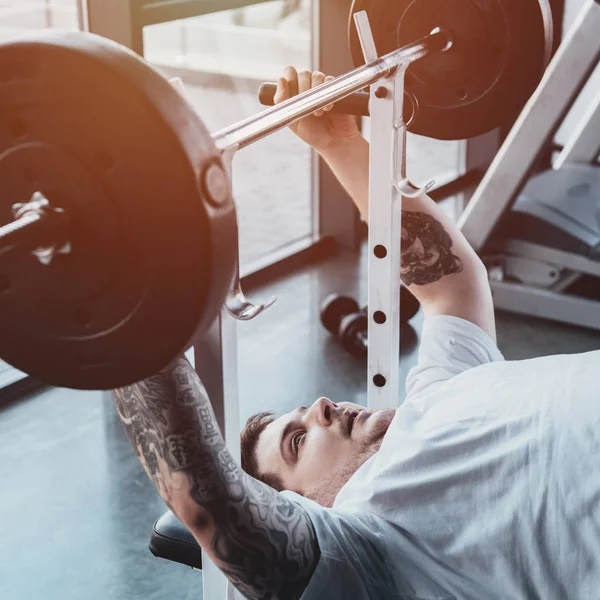 Sobrepeso hombre tatuado entrenamiento con barra de pesas en el gimnasio con luz solar - foto de stock