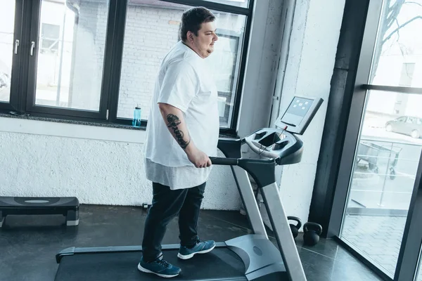 Overweight tattooed man in white t-shirt running on treadmill at gym — Stock Photo