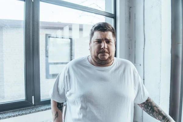Übergewichtiger, tätowierter Mann im weißen T-Shirt blickt in Turnhalle in die Kamera — Stockfoto
