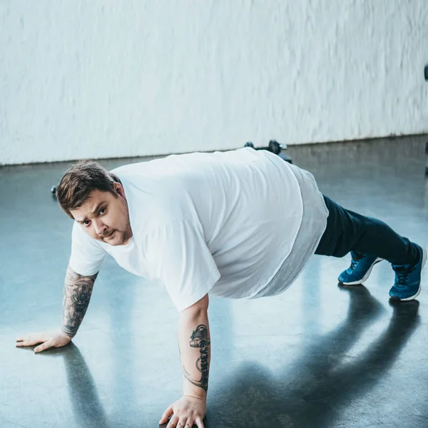 Hombre tatuado con sobrepeso mirando a la cámara mientras hace ejercicio push up en el centro deportivo - foto de stock
