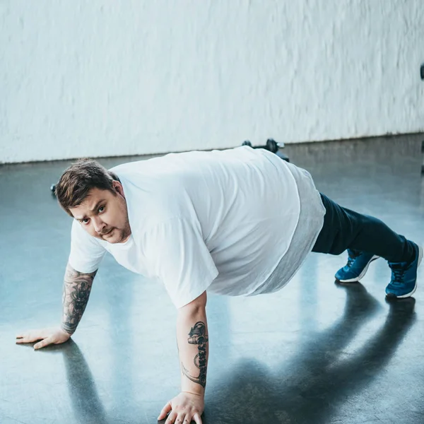 Übergewichtiger tätowierter Mann schaut in Kamera, während er Liegestützgymnastik im Sportzentrum macht — Stockfoto