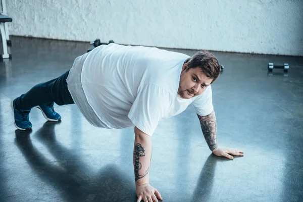 Hombre tatuado con sobrepeso mirando a la cámara mientras hace ejercicio push up en el centro deportivo - foto de stock