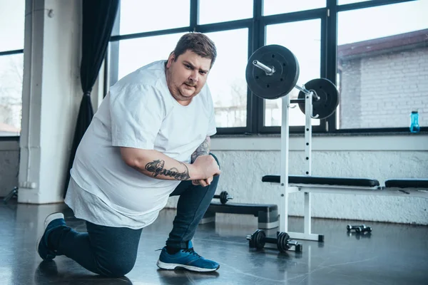 Übergewichtiger tätowierter Mann blickt in Kamera und macht Stretchübung im Fitnessstudio — Stockfoto