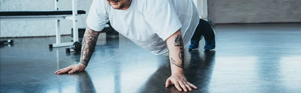 Tiro panorâmico de homem tatuado com sobrepeso fazendo push-up exercício no centro de esportes — Fotografia de Stock