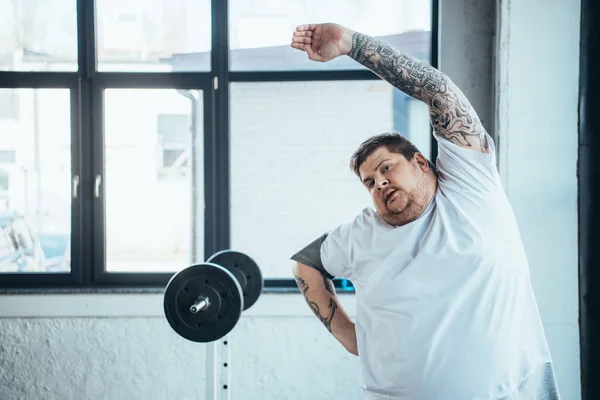 Übergewichtiger tätowierter Mann blickt in Kamera und macht Stretchübung im Fitnessstudio — Stockfoto
