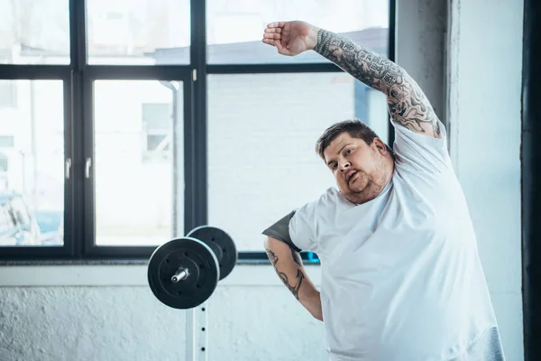 Hombre tatuado con sobrepeso mirando la cámara y haciendo ejercicio de estiramiento en el gimnasio - foto de stock