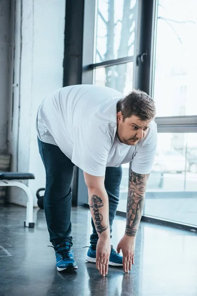 Hombre tatuado con sobrepeso en camiseta blanca haciendo ejercicio de estiramiento en el centro deportivo - foto de stock