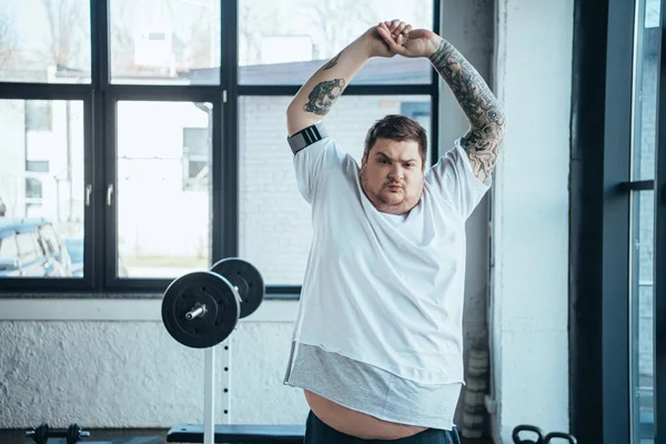 Hombre tatuado con sobrepeso mirando la cámara y haciendo ejercicio de estiramiento en el gimnasio - foto de stock