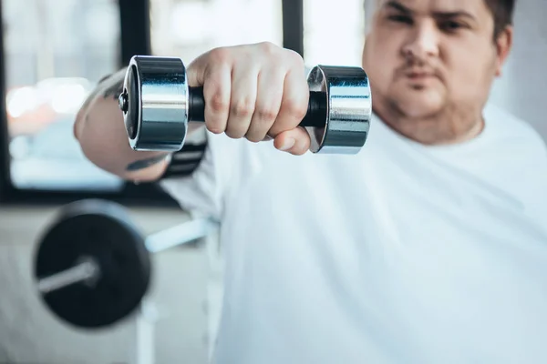 Обрезанный вид человека с избыточным весом, смотрящего на камеру во время тренировки с гантелями в тренажерном зале — стоковое фото