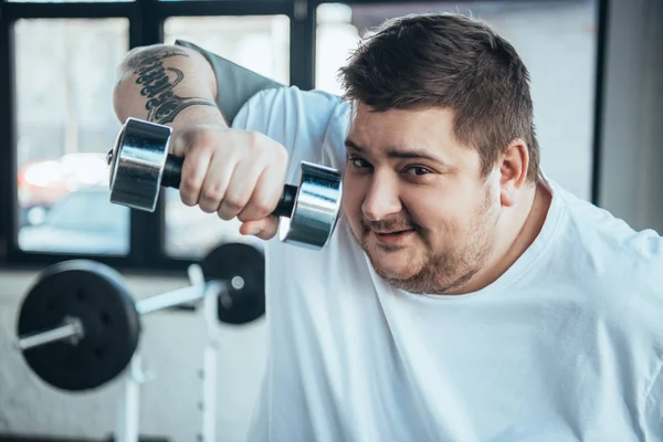 Smiling Overweight tattooed man Looking At Camera and exercising with dumbbell at sports center — Stock Photo