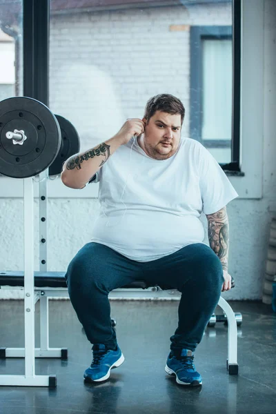 Sovrappeso tatuato uomo in cuffia seduto sulla panchina, guardando la fotocamera e ascoltando musica in palestra — Foto stock