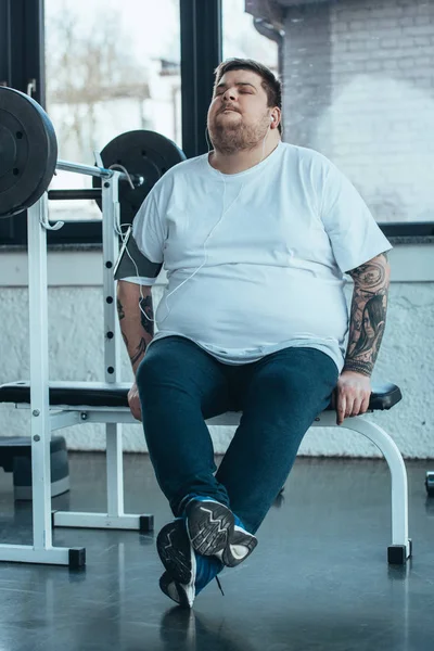 Overweight tattooed man in earphones sitting on bench and listening music at gym — Stock Photo