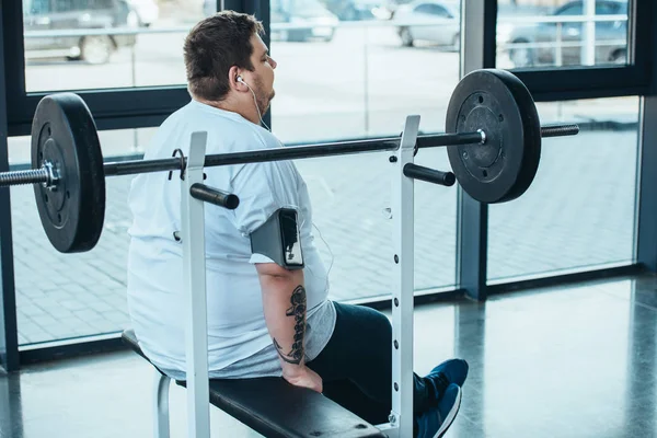 Übergewichtiger Mann mit Kopfhörer sitzt auf Bank und hört Musik im Fitnessstudio — Stockfoto