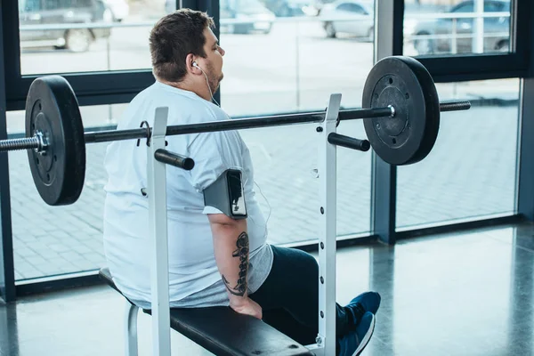 Hombre con sobrepeso en auriculares sentados en el banco y escuchando música en el gimnasio - foto de stock