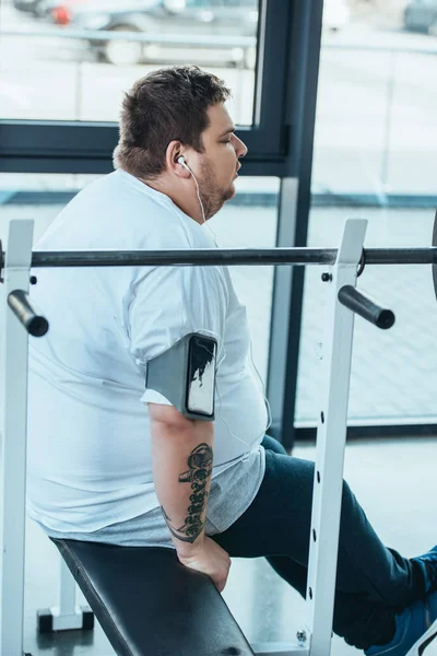 Hombre con sobrepeso en auriculares sentados en el banco y escuchando música en el gimnasio - foto de stock