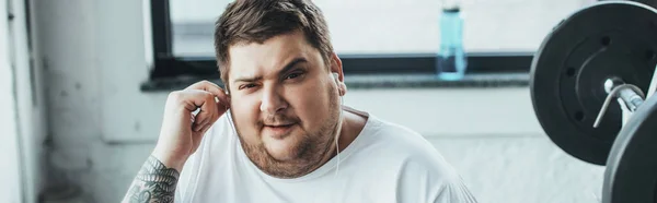 Panoramaaufnahme eines übergewichtigen Mannes, der im Fitnessstudio in die Kamera schaut, während er sich Kopfhörer aufsetzt — Stockfoto