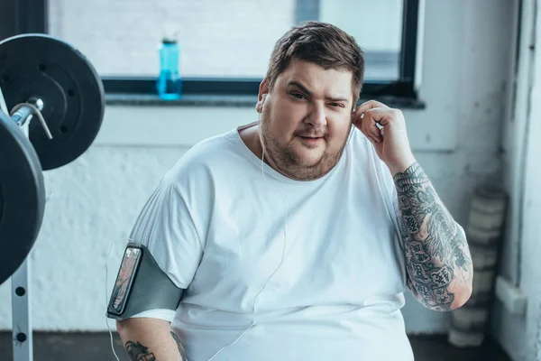 Hombre con sobrepeso en brazalete de teléfono inteligente mirando a la cámara mientras se pone los auriculares en el gimnasio - foto de stock