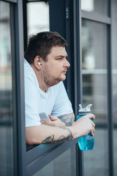 Overweight man with earphones and smartphone holding sport bottle and looking out through window at gym — Stock Photo