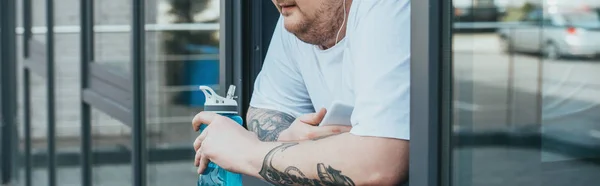 Panoramic shot of overweight man with earphones and smartphone holding sport bottle and looking out through window at gym — Stock Photo