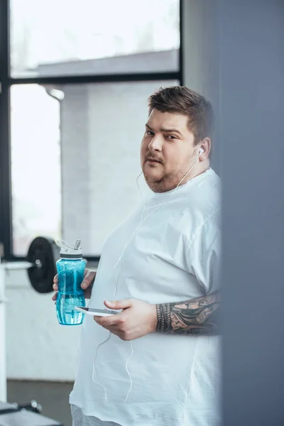 Hombre tatuado con sobrepeso sosteniendo la botella deportiva, mirando a la cámara y usando el teléfono inteligente en el gimnasio - foto de stock