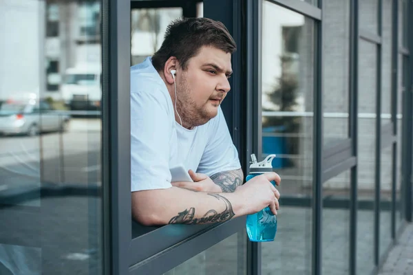 Overweight man with earphones and smartphone holding sport bottle and looking out through window at gym — Stock Photo