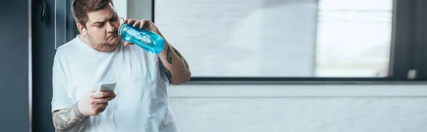 Панорамный снимок человека с татуировкой с избыточным весом, использующего смартфон и питьевую воду из спортивной бутылки в спортзале — стоковое фото