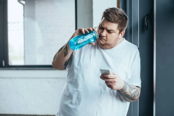 Hombre tatuado con sobrepeso usando teléfono inteligente y agua potable de la botella de deporte en el gimnasio - foto de stock