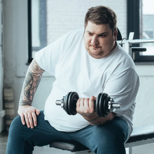 Overweight tattooed man sitting and exercising with dumbbell at sports center — Stock Photo