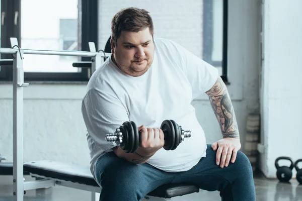 Overweight tattooed man sitting and training with dumbbell at sports center — Stock Photo