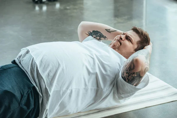 Übergewichtiger tätowierter Mann liegt auf Fitnessmatte und macht Aufsitzgymnastik im Sportzentrum — Stockfoto