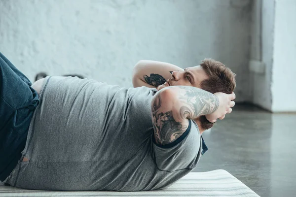 Übergewichtiger tätowierter Mann liegt auf Fitnessmatte und macht Aufsitzgymnastik im Sportzentrum — Stockfoto