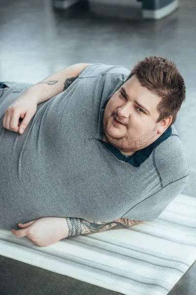 Hombre tatuado con sobrepeso en camiseta gris mirando a la cámara mientras está acostado en la colchoneta de fitness en el centro deportivo - foto de stock