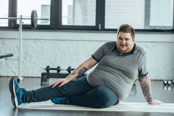 Uomo tatuato sovrappeso guardando la fotocamera, sorridente e seduto sul tappeto fitness al centro sportivo — Foto stock