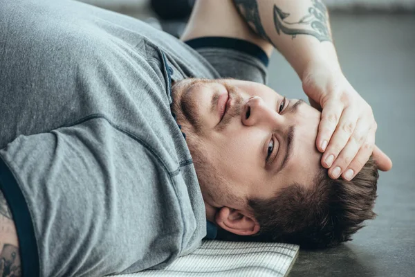 Erschöpfter, übergewichtiger, tätowierter Mann im grauen T-Shirt blickt in die Kamera, während er im Sportzentrum auf einer Fitnessmatte liegt — Stockfoto