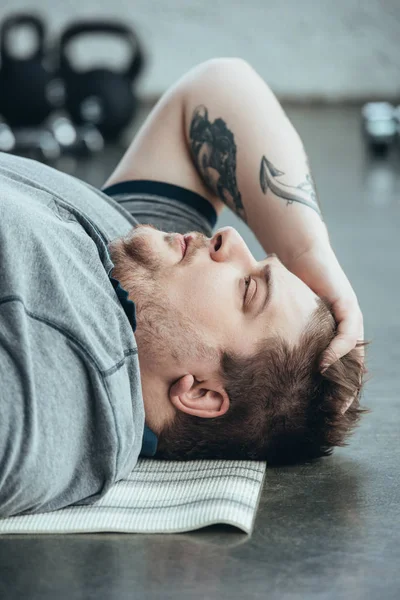 Виснажений татуйований чоловік з надмірною вагою у сірій футболці, що лежить на фітнес-матусі у спортивному центрі — стокове фото