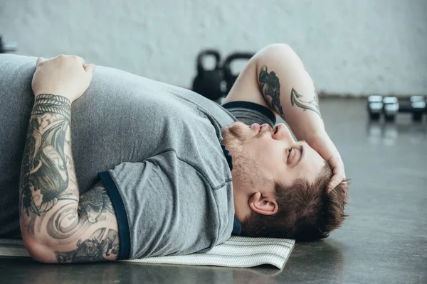 Tired overweight tattooed man in grey t-shirt lying on fitness mat at sports center — Stock Photo