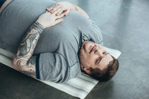 Müder, übergewichtiger, tätowierter Mann im grauen T-Shirt blickt in die Kamera, während er im Sportzentrum auf einer Fitnessmatte liegt — Stockfoto