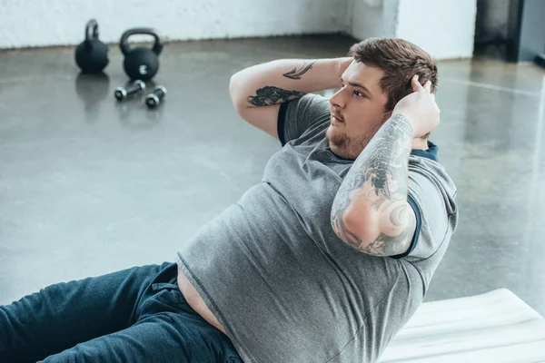 Sobrepeso tatuado homem fazendo sentar-se exercício no centro de esportes — Fotografia de Stock